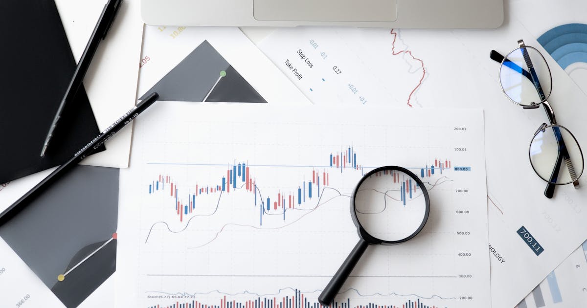 Financial analysis documents and stock market data spread across a desk with magnifying glass